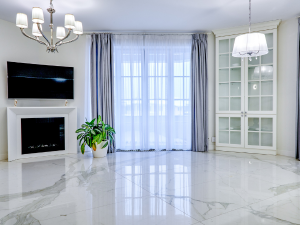 Bright living room with marble floors, a fireplace and wall-mounted TV, a green potted plant, and a glass-front cabinet by a large window with sheer curtains.